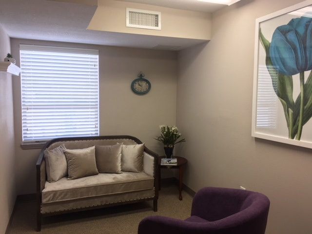 Image of mental health counseling office with a cream velvet couch, accompanied by a purple chair and wood side table. The walls have a large picture of a blue and green tulip, teal clock, and hanging shelf with a plant. A window is also present.